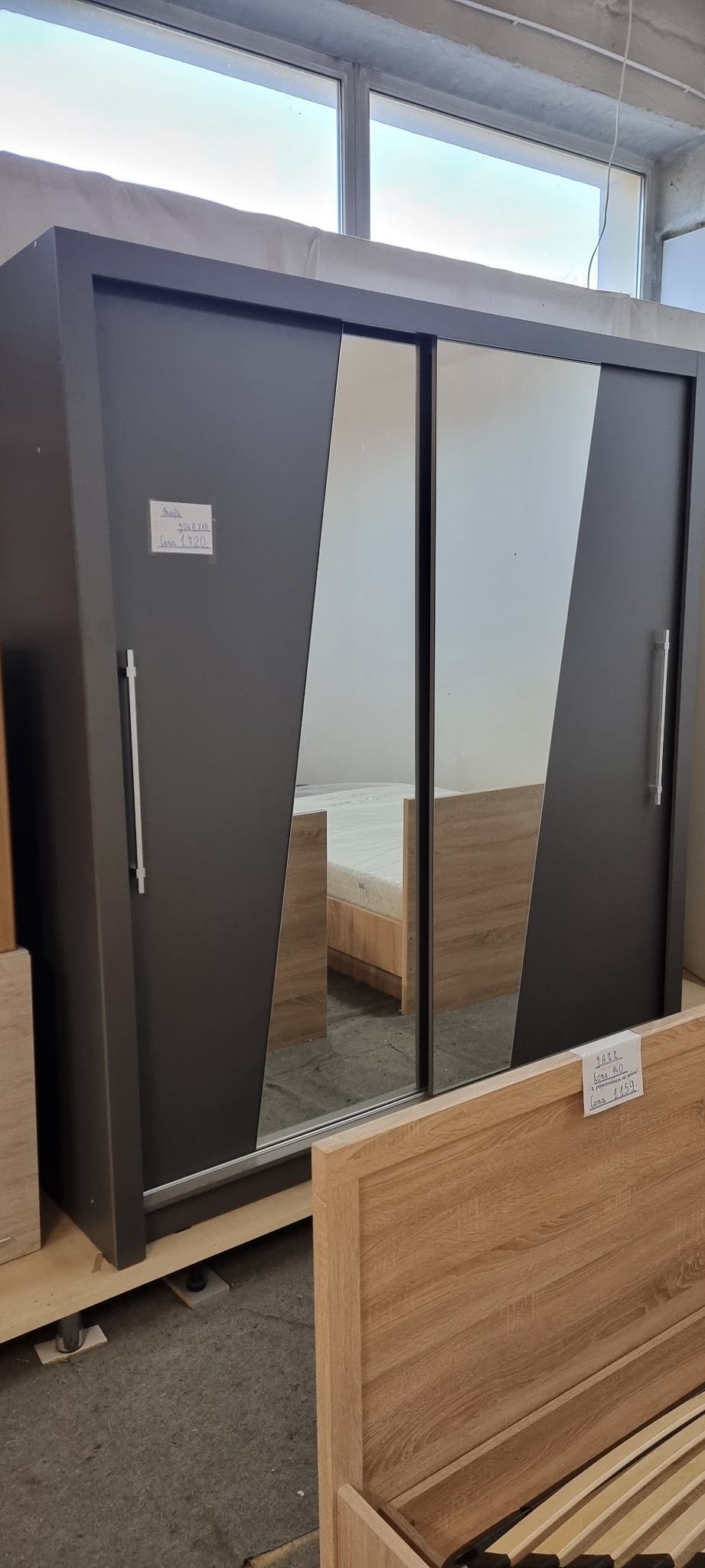 Ciemnoszara szafa przesuwna z lustrami o ukośnych krawędziach, widoczna w surowym wnętrzu magazynu z oknami, obok elementy drewnianego łóżka, na szafie naklejka z ceną.
