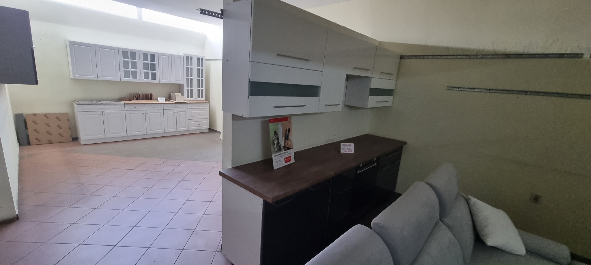 Wystawa mebli kuchennych: białe szafki wiszące z szarymi akcentami, ciemny blat, widoczna częściowo szara sofa w tle, oraz dalszy plan z białymi szafkami kuchennymi, prezentowane w przestronnym...