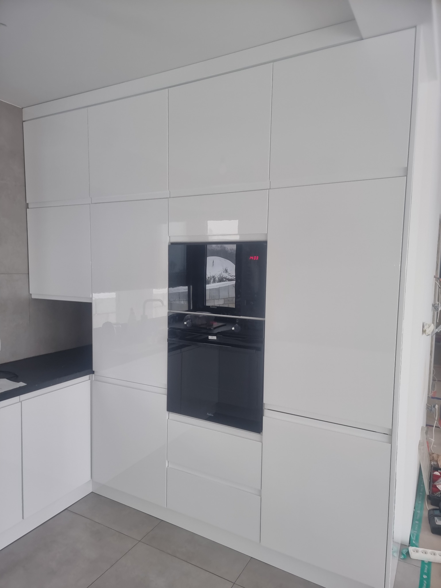 Minimalistyczna, biała zabudowa kuchenna z błyszczącymi frontami, wbudowany piekarnik z czerwonym wyświetlaczem godziny 14:03, szare płytki podłogowe, widoczne elementy instalacji elektrycznej w rogu.