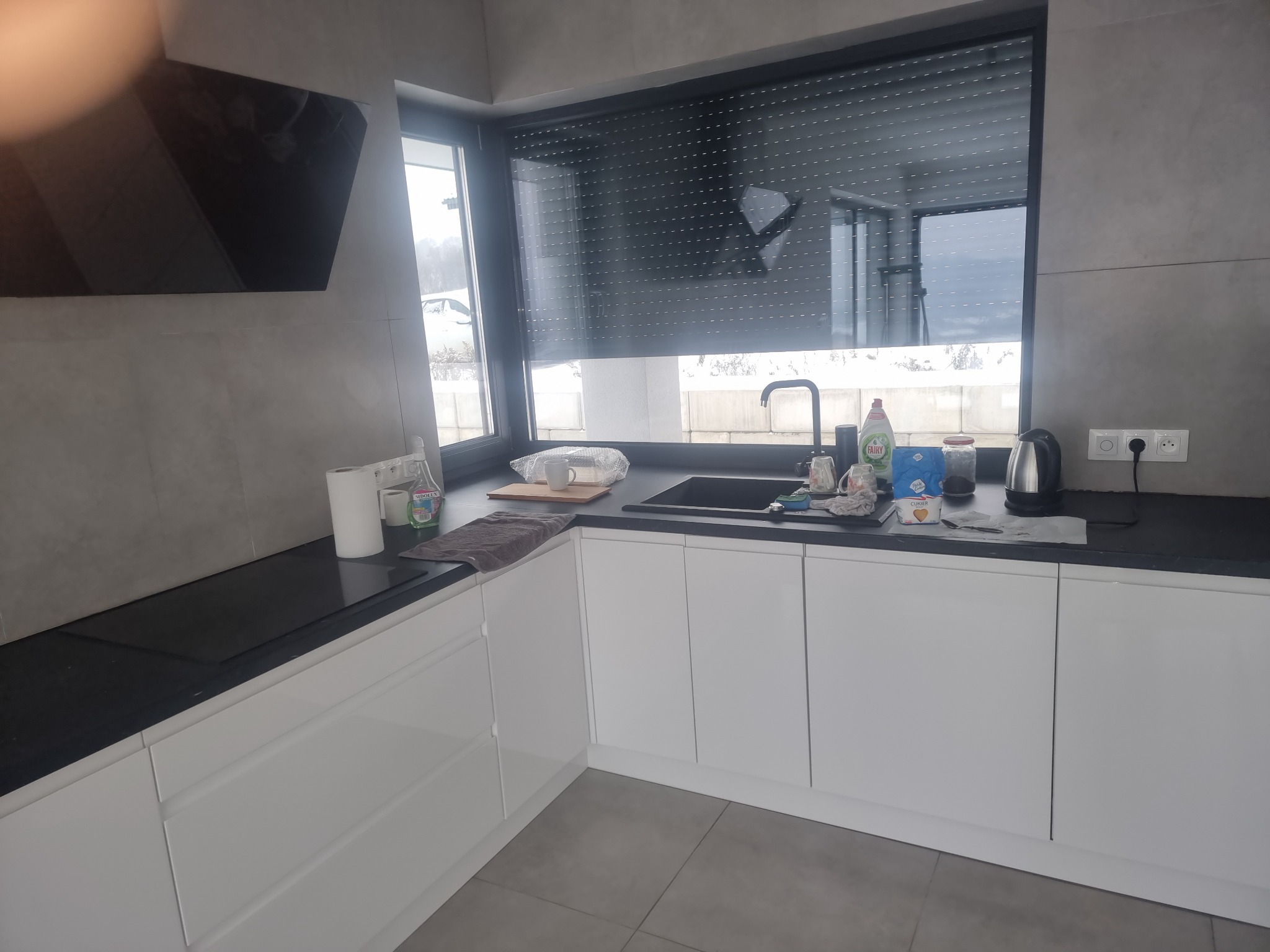 Nowoczesna kuchnia z białymi szafkami, czarnym blatem i zlewozmywakiem, widok na okno z roletami zewnętrznymi, na blacie widoczne akcesoria kuchenne i płyn do naczyń