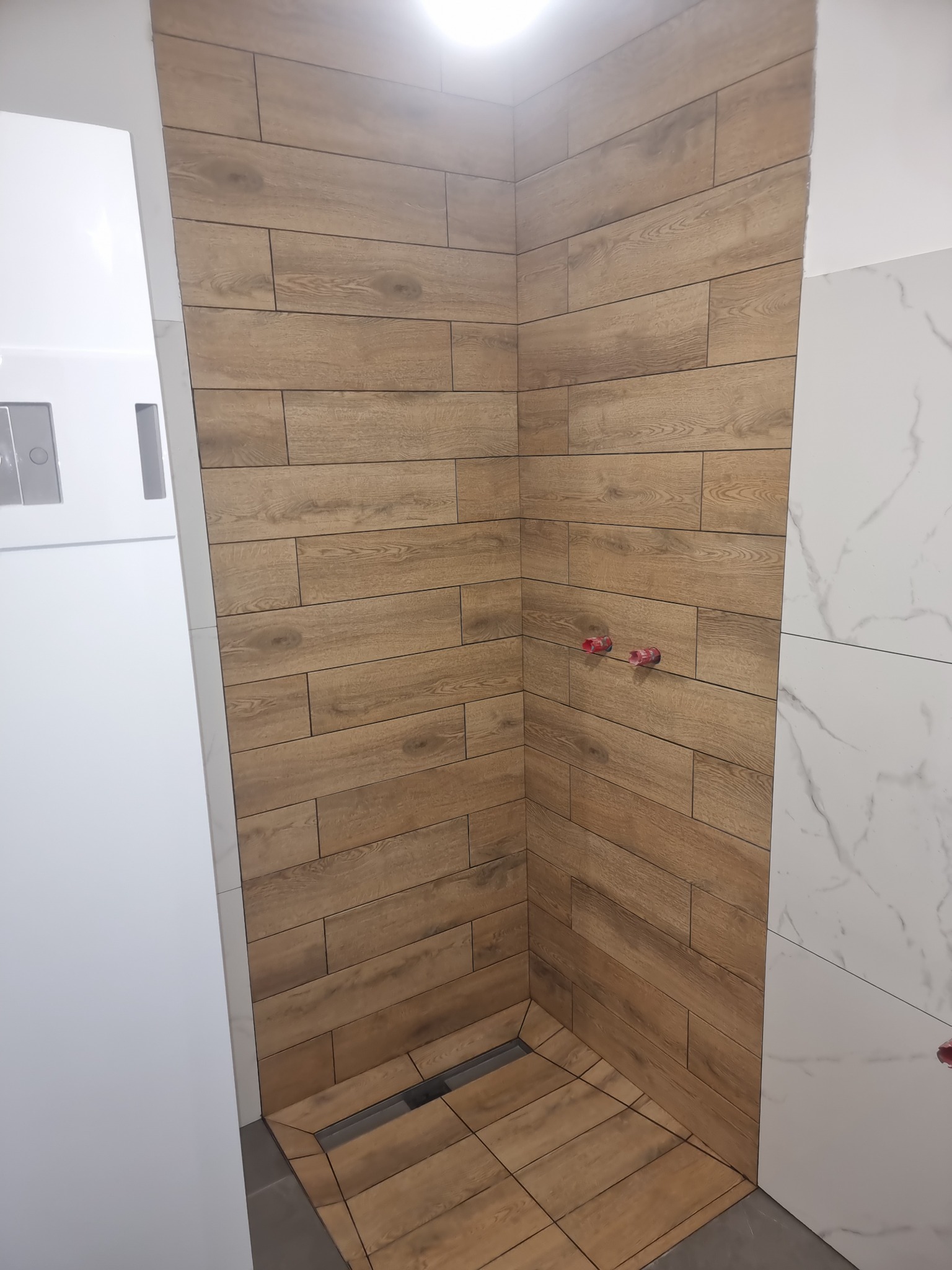 Wykończony fragment kabiny prysznicowej z drewnopodobnymi płytkami na ścianach i podłodze, widoczne przyłącza wodne i odpływ liniowy.