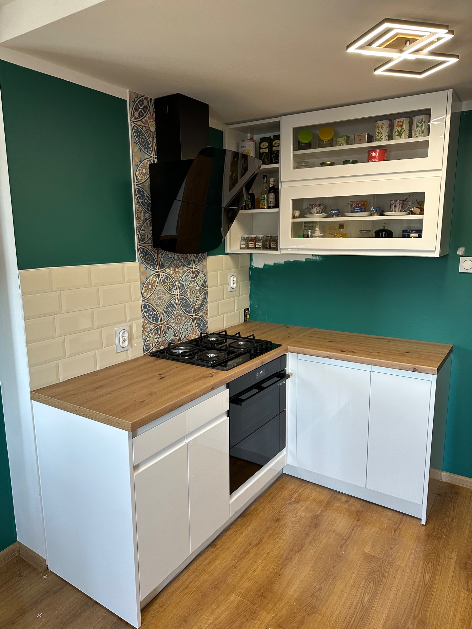 Nowoczesna kuchnia z białymi szafkami, drewnianym blatem i zieloną ścianą, urozmaicona płytkami dekoracyjnymi nad kuchenką gazową oraz przeszklonymi szafkami wiszącymi z widoczną zawartością.