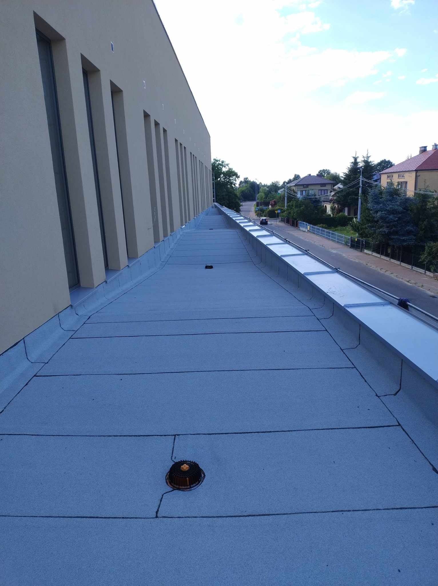 Płaski dach pokryty szarą membraną bitumiczną z widocznym kominkiem wentylacyjnym i obróbką blacharską, budynek z długimi oknami w tle.