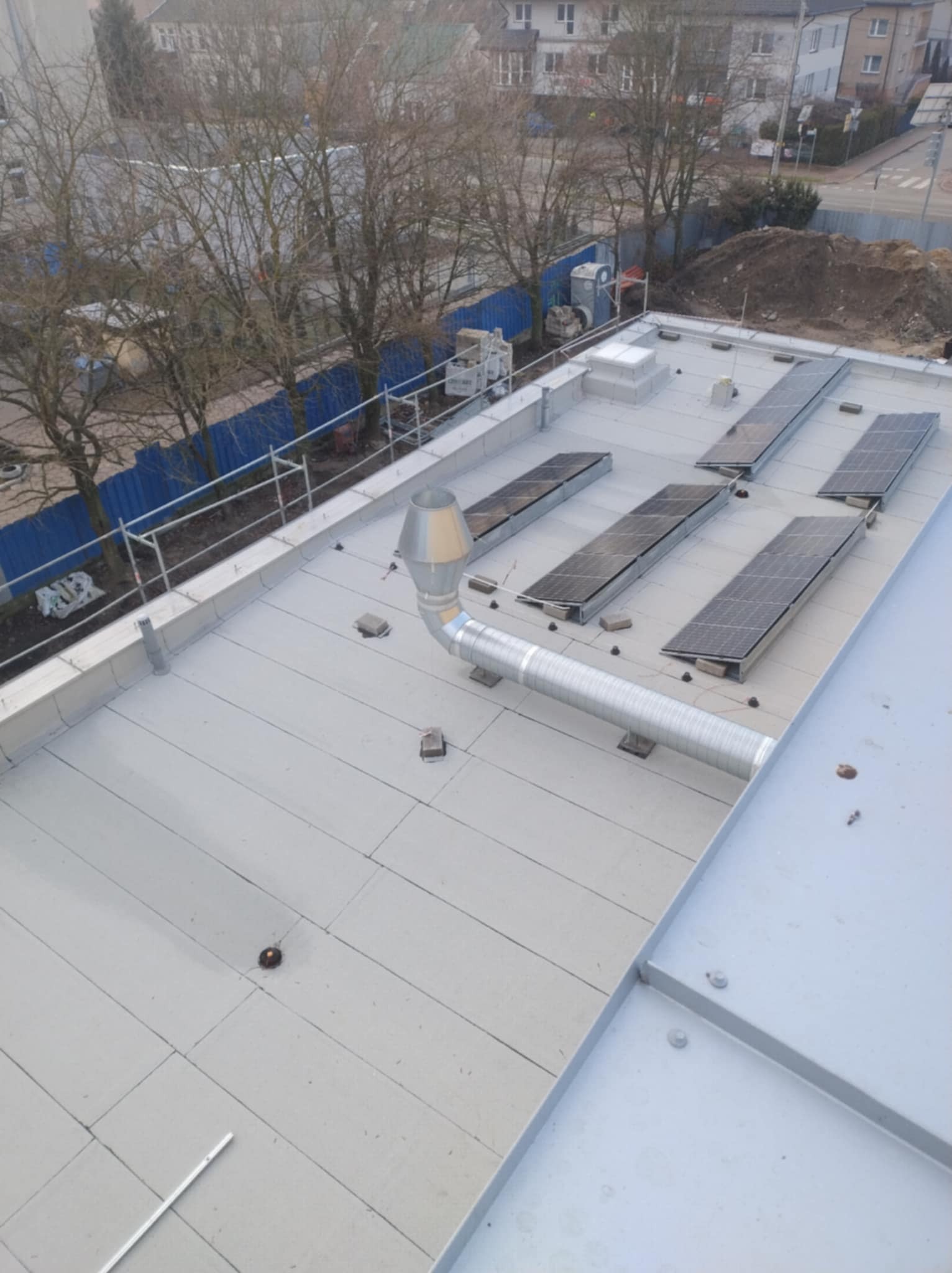 Płaski, jasnoszary dach z panelami słonecznymi, kominem wentylacyjnym i systemem asekuracyjnym, widok z góry na tle zabudowy miejskiej.