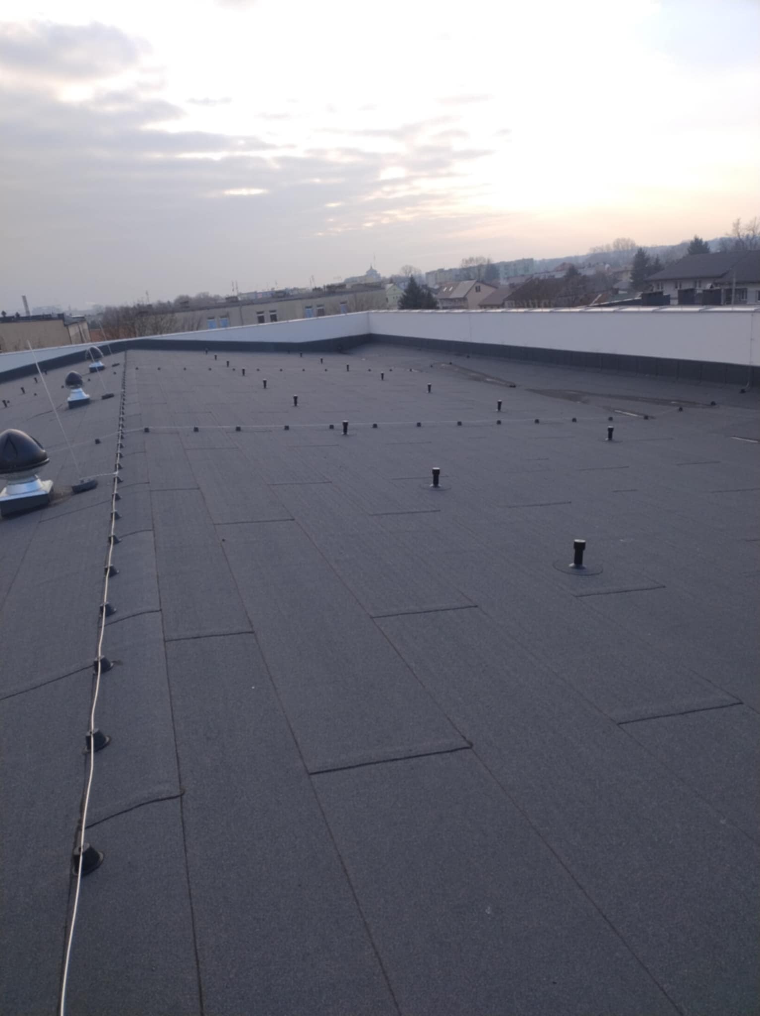 Czarny, płaski dach pokryty papą termozgrzewalną z widocznymi odpowietrznikami i systemem odgromowym, na tle zabudowań miejskich i pochmurnego nieba.