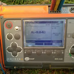 Miernik Sonel MPI-520 na trawie, wyświetlający wynik pomiaru rezystancji uziemienia Re=9,04Ω, z podłączonymi przewodami pomiarowymi. Widoczna dłoń osoby obsługującej urządzenie.
