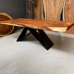 Stół z blatem z litego drewna egzotycznego o nieregularnych krawędziach, wsparty na czarnych, metalowych nogach o geometrycznym kształcie. W tle widoczne drewniane plastry i deski.