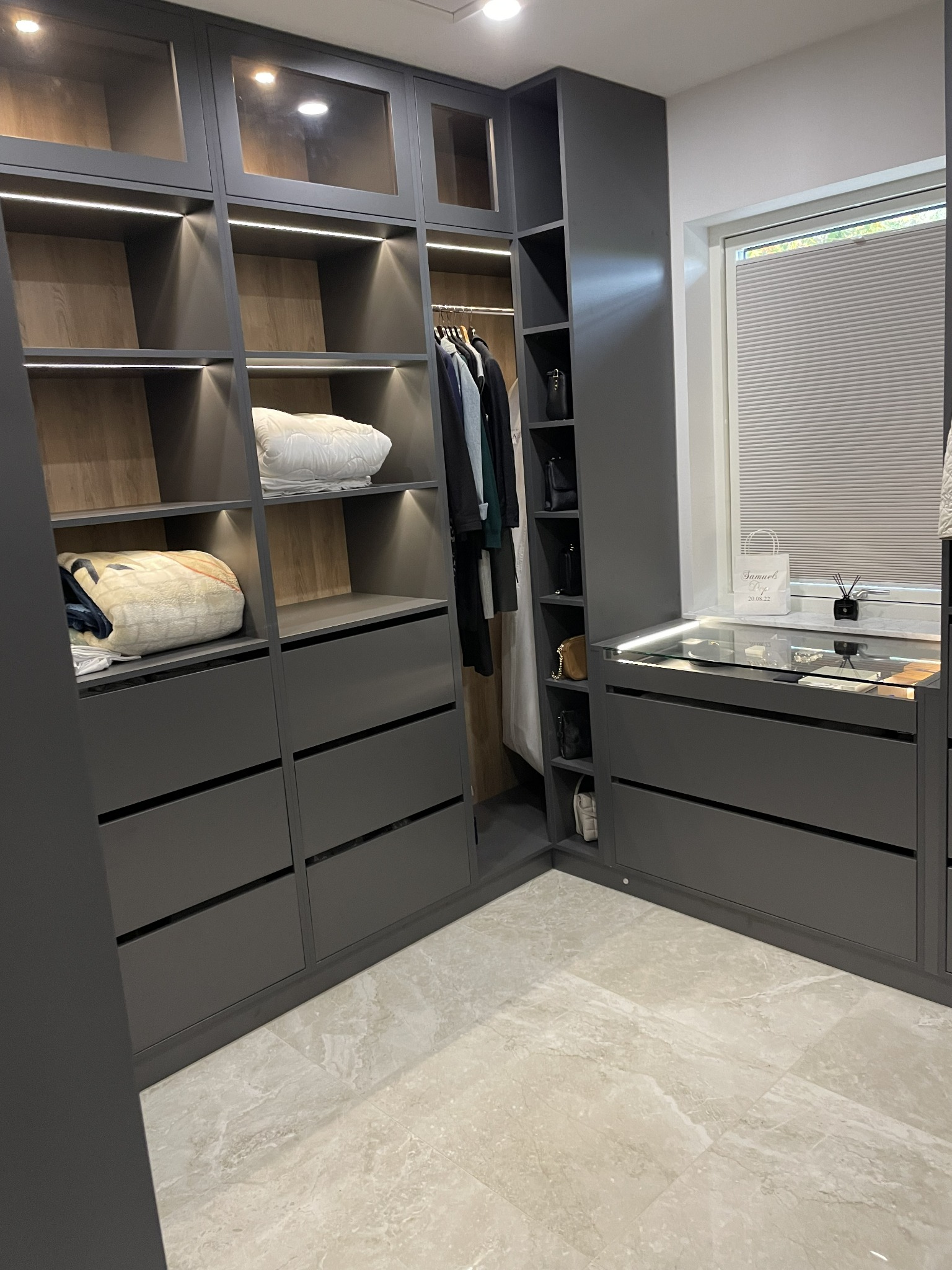 Szara garderoba na wymiar z oświetleniem LED, częściowo przeszklonymi drzwiami i półkami, widoczne ubrania i akcesoria, marmurowe płytki podłogowe.