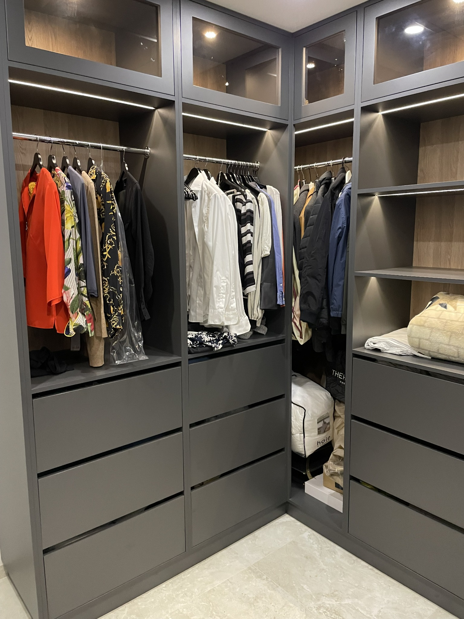 Szara, narożna szafa na wymiar z szufladami i przeszklonymi górnymi szafkami, oświetlona listwami LED, z ubraniami na wieszakach i torbami do przechowywania na półkach.
