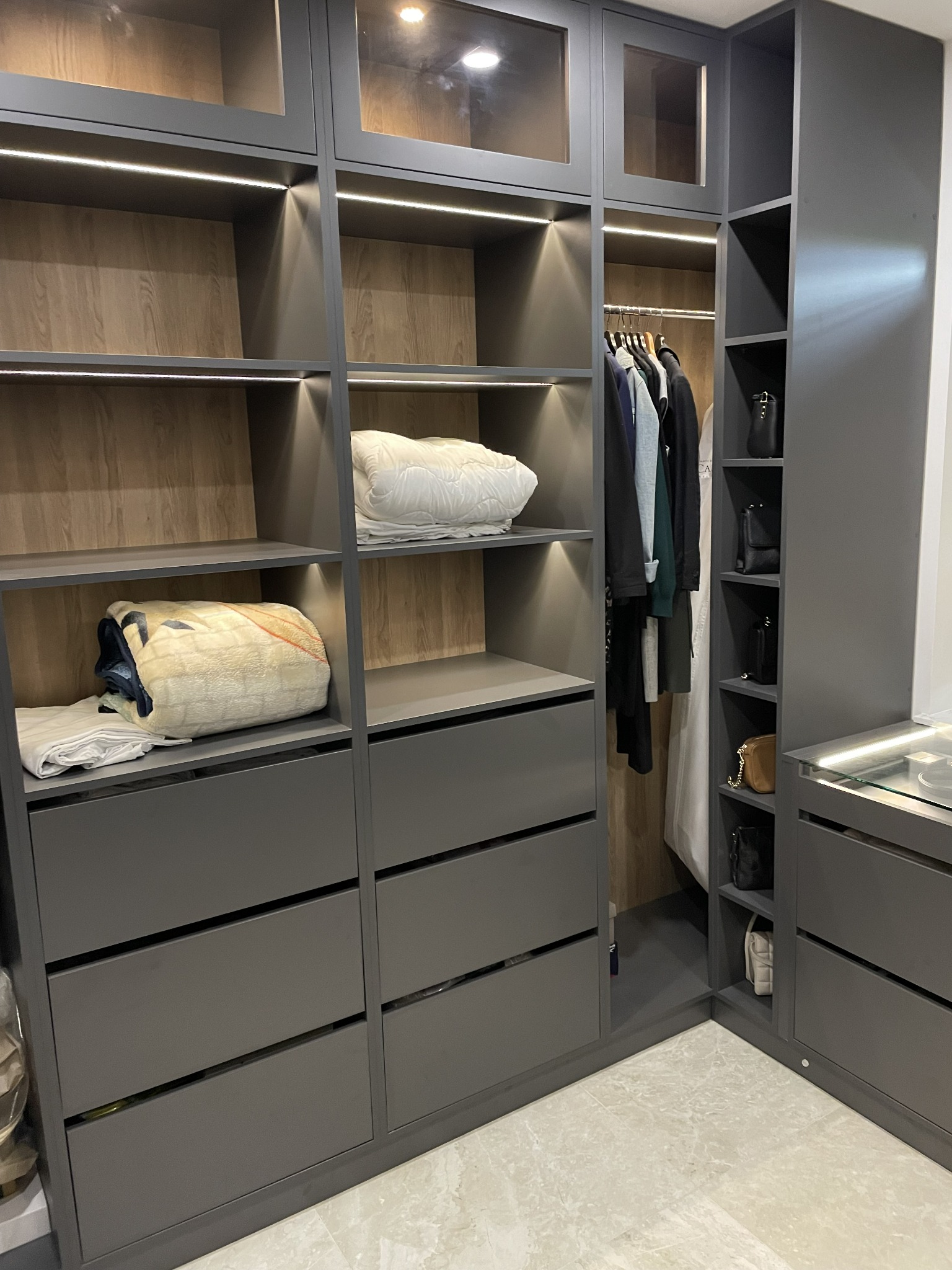 Szara szafa na wymiar z oświetleniem LED, półkami na ubrania i szufladami, wypełniona złożonymi tekstyliami i wiszącymi ubraniami.