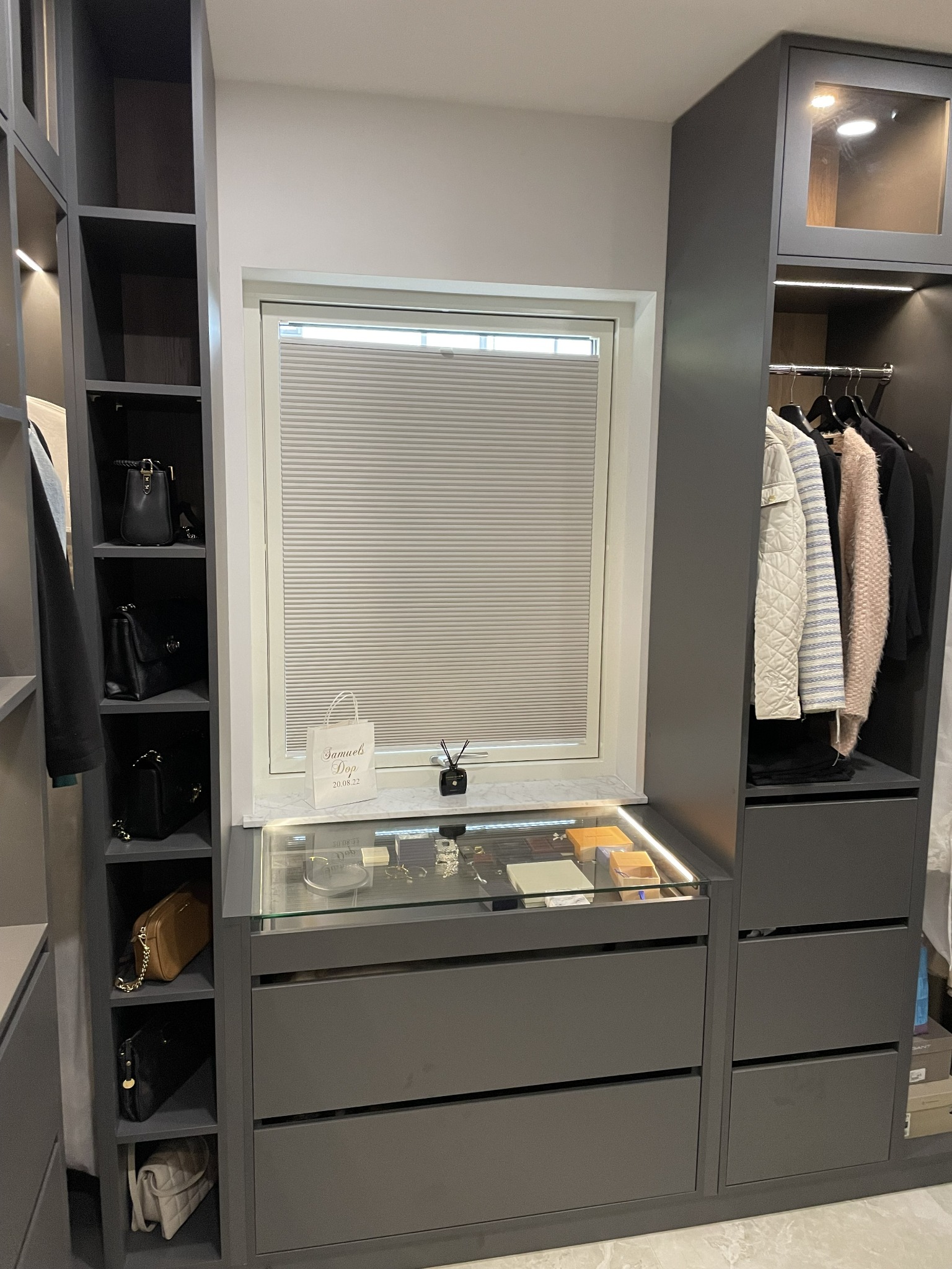 Szara szafa na wymiar z szufladami, przeszkloną witryną na biżuterię, otwartymi półkami na torebki i miejscem na ubrania wiszące, oświetlona punktowo, z żaluzjami okiennymi w tle.