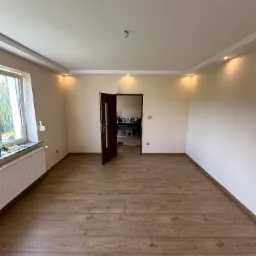 Pusty pokój po remoncie z drewnianą podłogą, świeżo pomalowanymi ścianami w odcieniach beżu i zamontowanym oświetleniem punktowym w suficie podwieszanym, widoczne otwarte drzwi wewnętrzne.