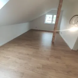 Poddasze po remoncie z drewnianą podłogą w panelach, białe ściany i drewniany słup konstrukcyjny, widok na okno dachowe.