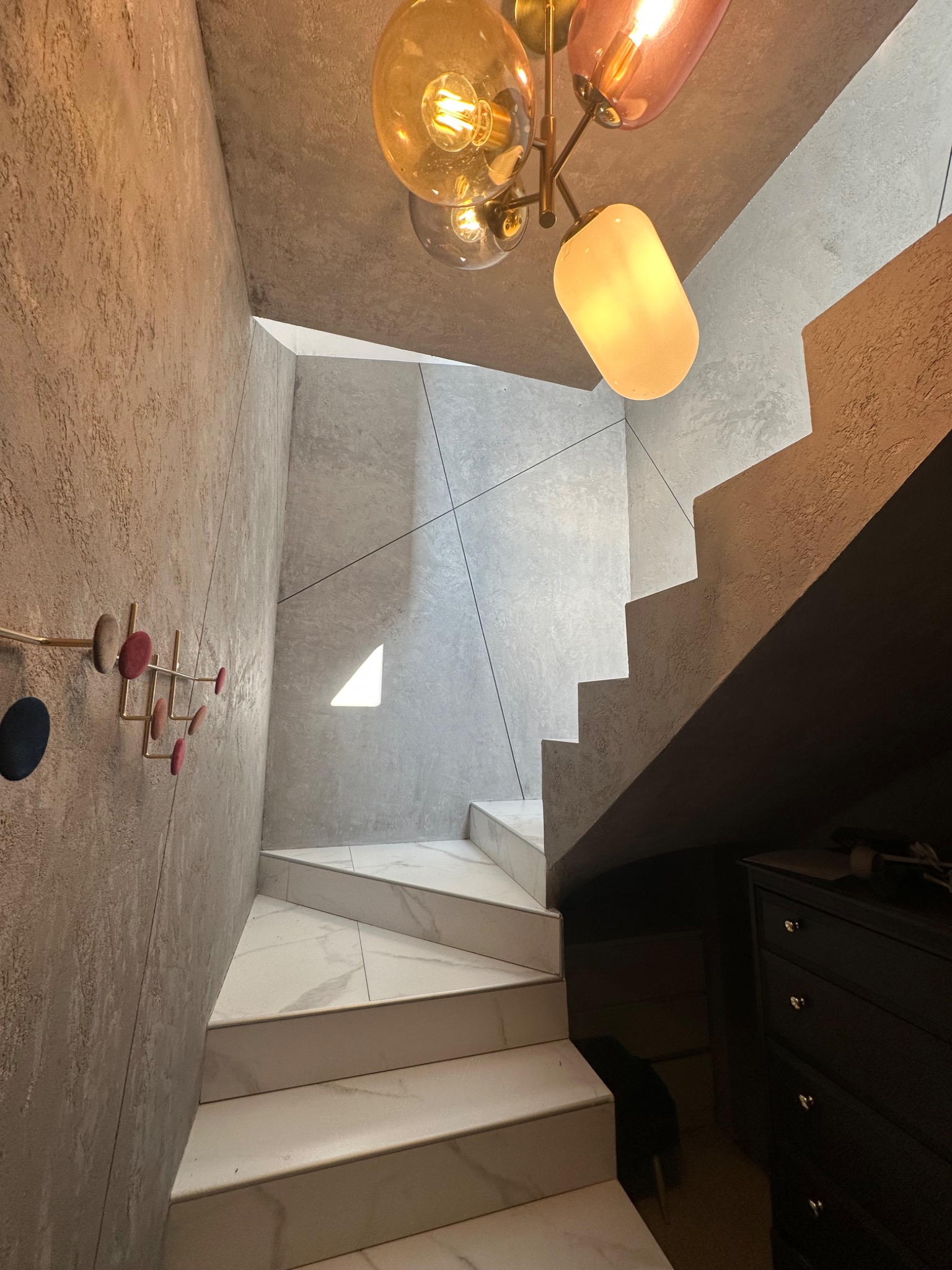 Nowoczesna klatka schodowa z marmurowymi stopniami, szarą teksturowaną ścianą z wieszakiem w stylu retro i designerską lampą wiszącą z kolorowymi kloszami.