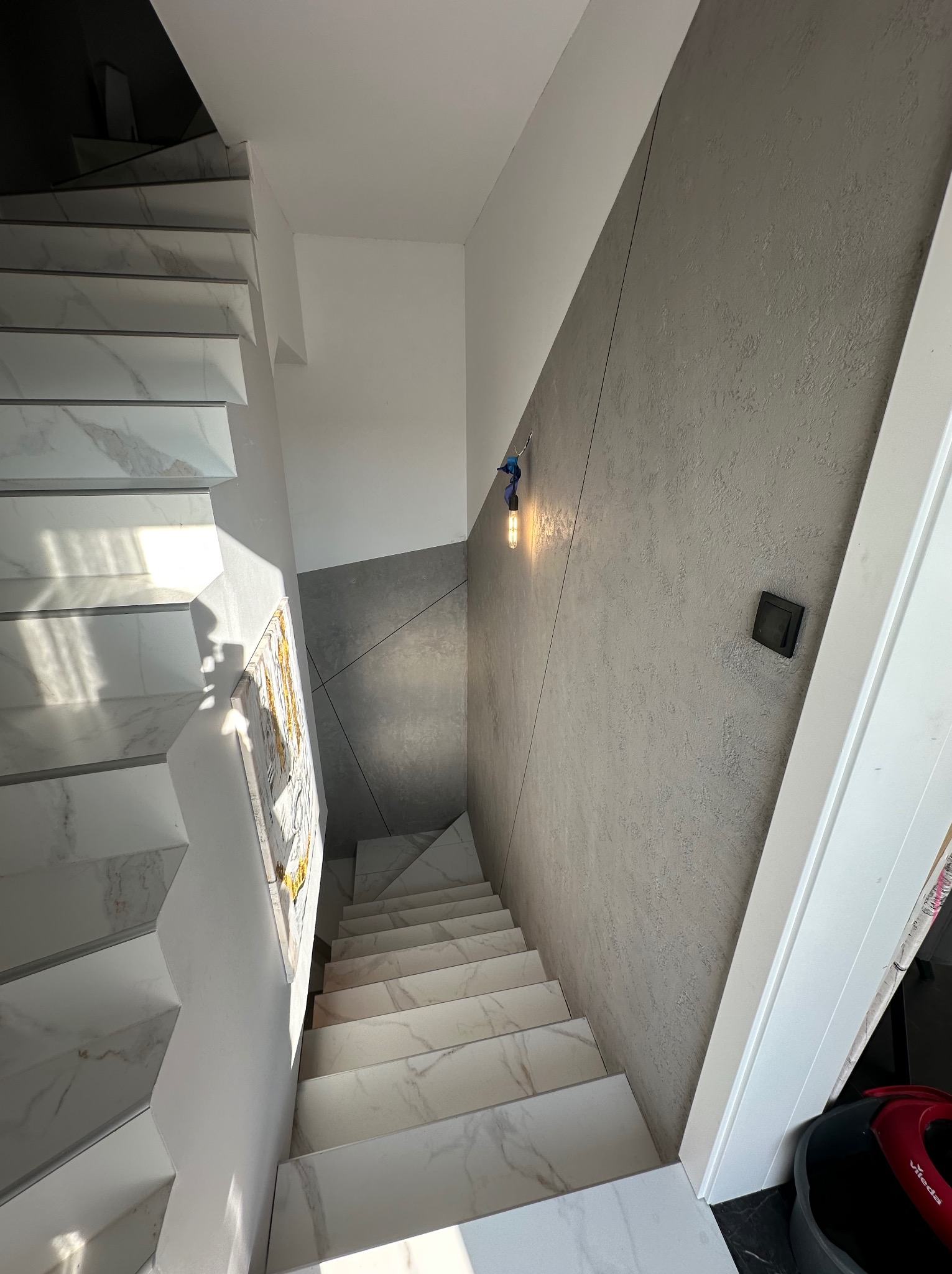 Wnętrze klatki schodowej z marmurowymi schodami, szarą ścianą z fakturą i dekoracyjną żarówką. Białe ściany i listwy wykończeniowe. Na podeście widoczny obraz z motywem złotych liści.