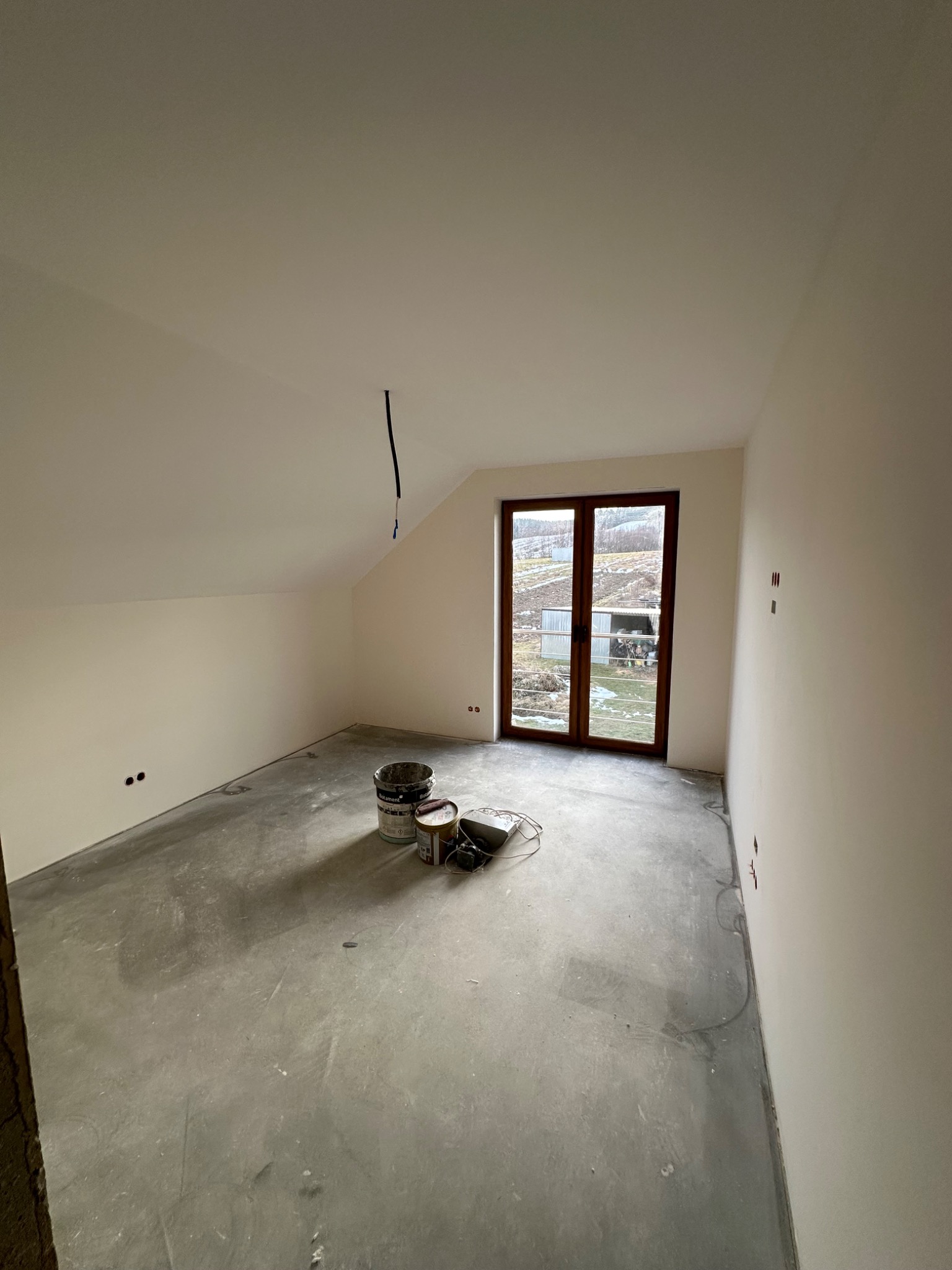 Poddasze w trakcie prac wykończeniowych: świeżo pomalowane ściany w odcieniach bieli, surowa betonowa podłoga, widoczne puszki z farbą i narzędzia malarskie, okno balkonowe z widokiem na zewnątrz.