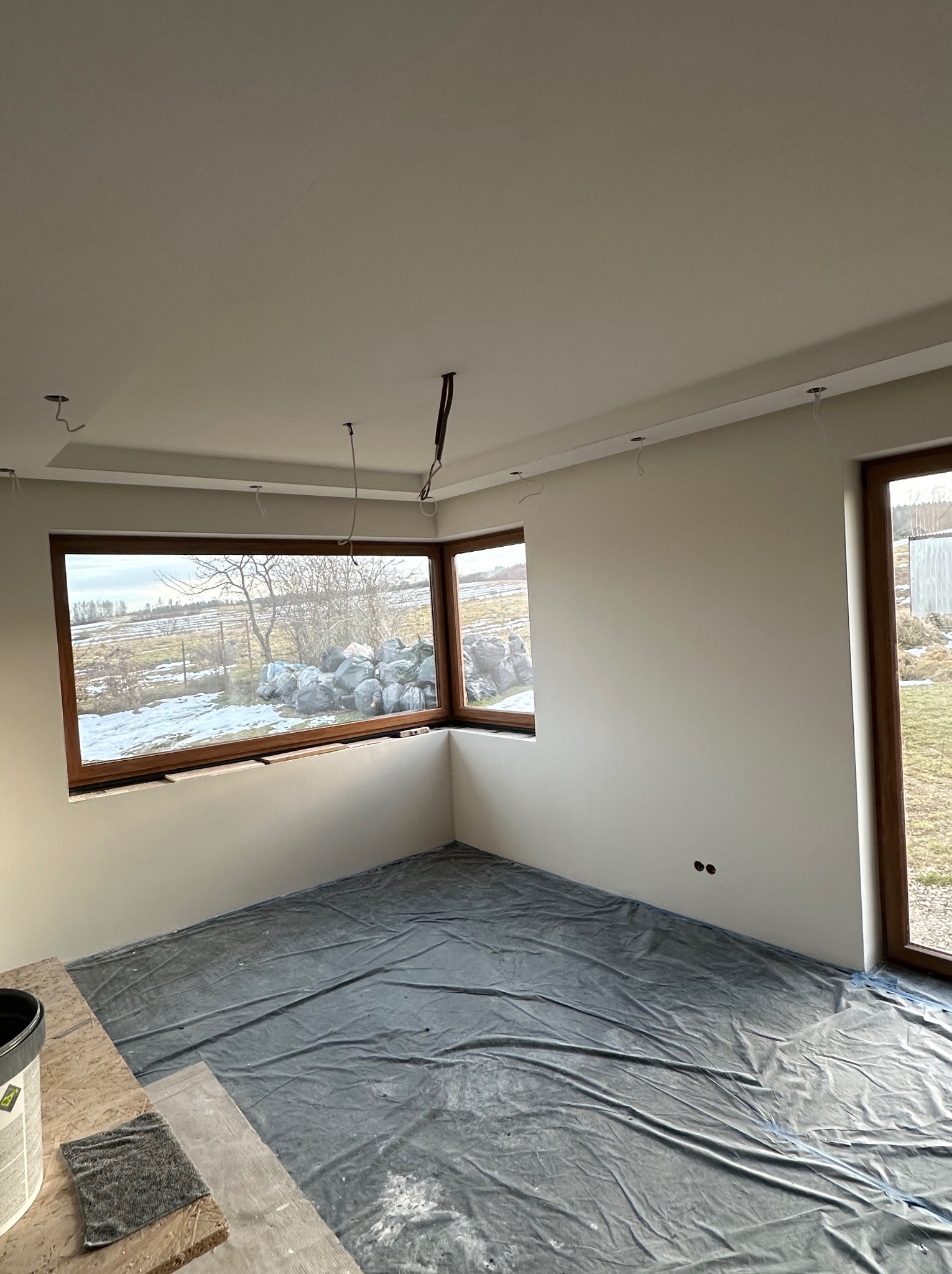 Pokój w trakcie prac wykończeniowych: świeżo pomalowane ściany na biało, zabezpieczona folią podłoga, surowe drewniane okna i wystające kable od oświetlenia sufitowego.