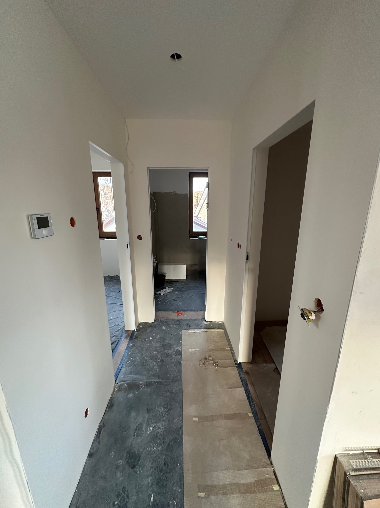 Korytarz w trakcie remontu: świeżo pomalowane ściany na biało, widoczne otwory na gniazdka, zabezpieczona podłoga i widok na sąsiednie pomieszczenia z oknami.