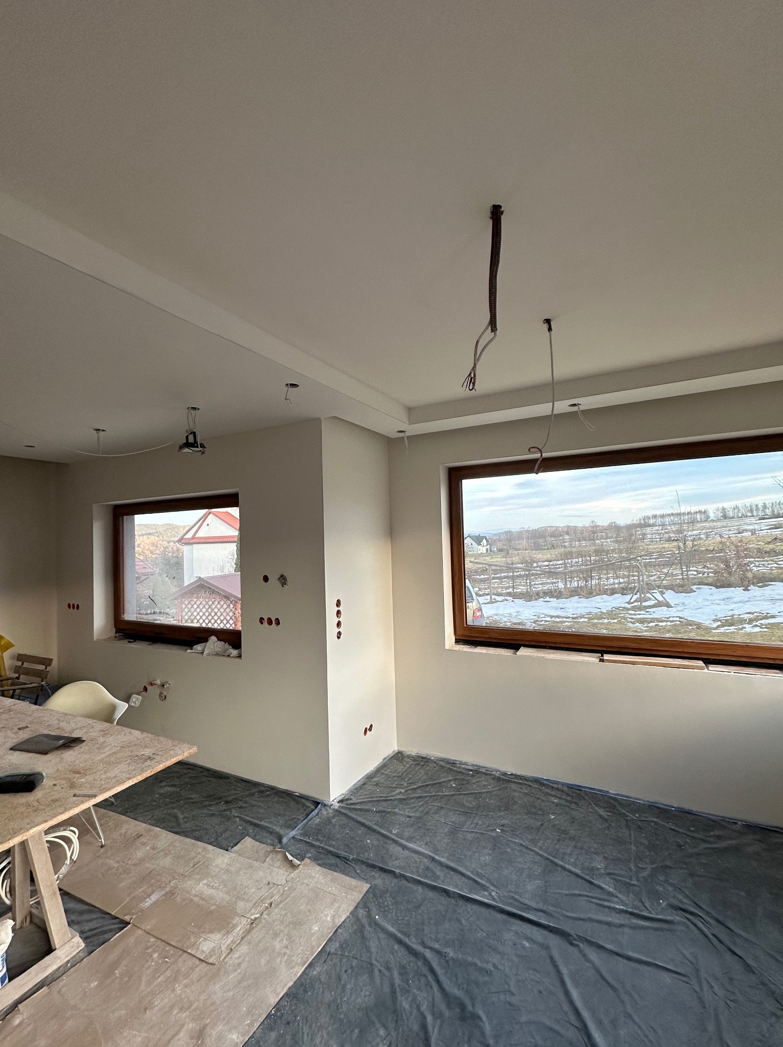 Wnętrze w trakcie prac wykończeniowych: świeżo pomalowane ściany w neutralnym odcieniu, zabezpieczona folią podłoga, widoczne otwory na gniazdka elektryczne i wystające kable oświetleniowe z sufitu...