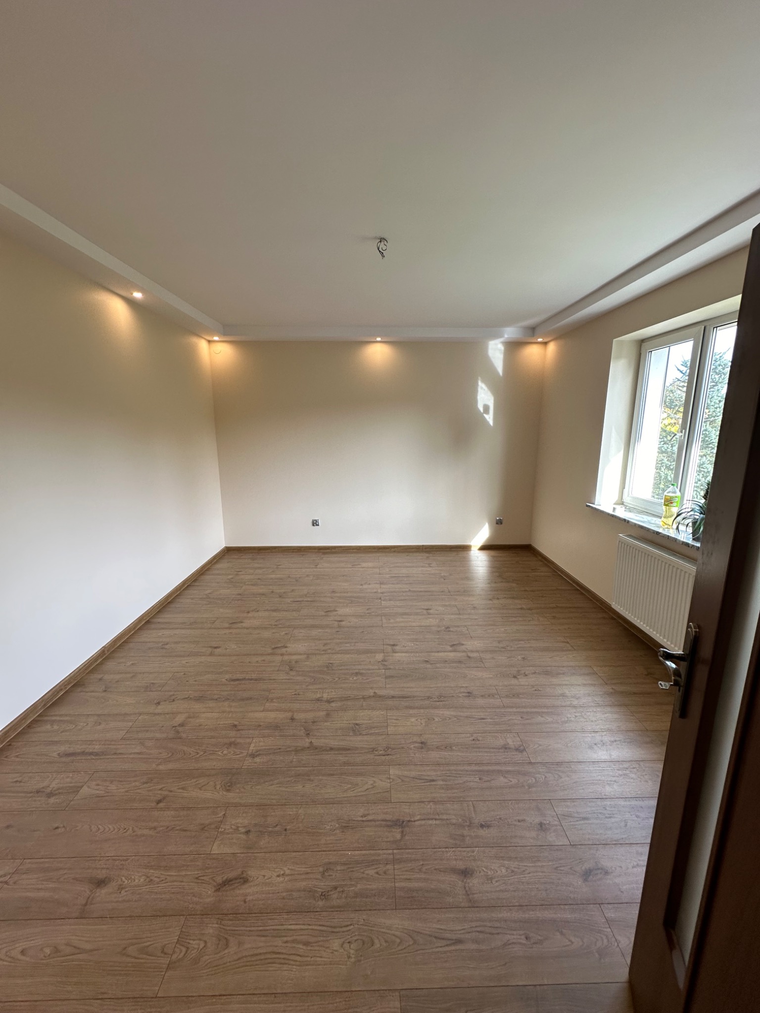 Pusty pokój po remoncie z panelami podłogowymi w odcieniach brązu, beżowymi ścianami i oświetleniem punktowym w suficie podwieszanym, widok na okno z grzejnikiem.