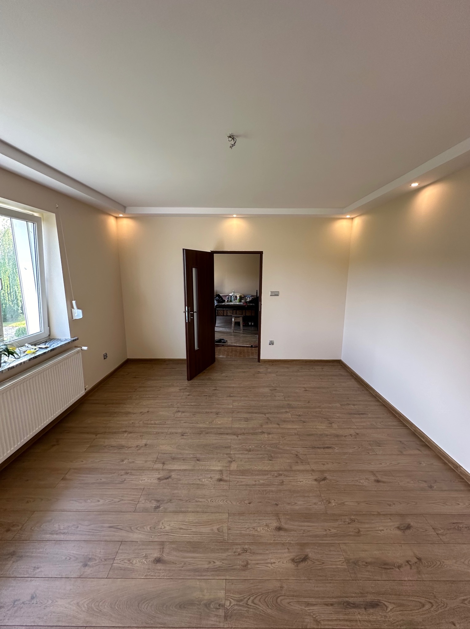 Pusty pokój po remoncie z drewnianą podłogą, świeżo pomalowanymi ścianami w odcieniach beżu i zamontowanym oświetleniem punktowym w suficie podwieszanym, widoczne otwarte drzwi wewnętrzne.