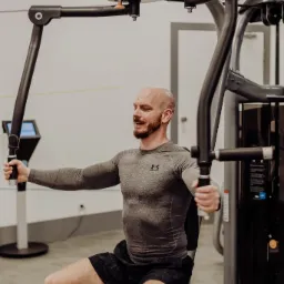 Mężczyzna z brodą ćwiczy na maszynie do rozpiętek w siłowni, ubrany w szarą koszulkę z długim rękawem i czarne spodenki, z widocznym logo marki sportowej.