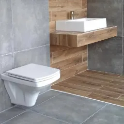 Nowoczesna łazienka z wiszącą toaletą, prostokątną umywalką na drewnianym blacie i szarymi płytkami imitującymi beton na ścianach, kontrastującymi z brązowymi płytkami imitującymi drewno...