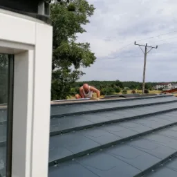 Pracownik w białym podkoszulku instaluje grafitowe panele dachowe, widok z okna na wiejski krajobraz i słup elektryczny.