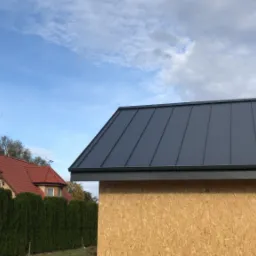 Nowy, grafitowy dach z blachy na drewnianej konstrukcji, w tle dom z czerwoną dachówką i zielony żywopłot, błękitne niebo z chmurami.