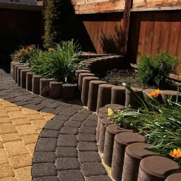 Ogród z dwukolorową ścieżką z kostki, obrzeża z betonowych palików, drewniane ogrodzenie w tle.