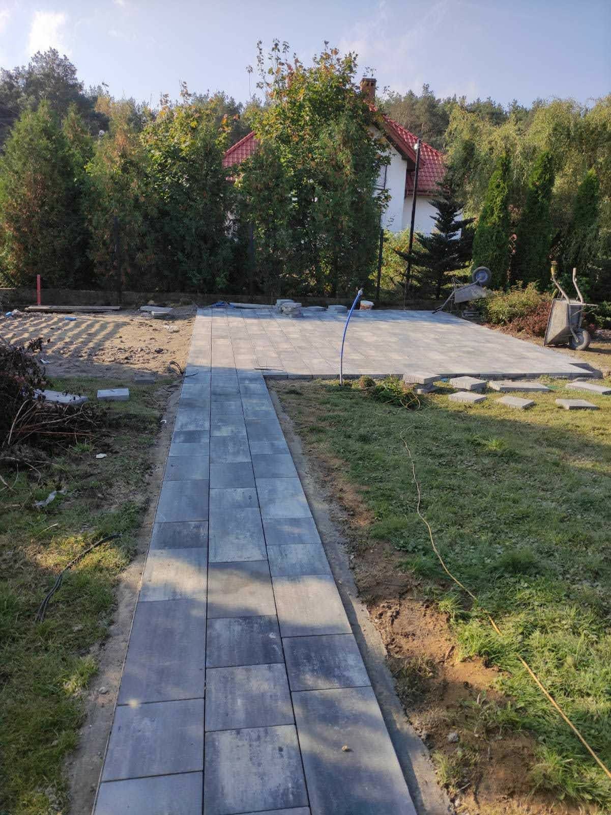 Fragment ścieżki z szarych płyt betonowych prowadzącej do częściowo ułożonego placu z kostki, tło z zielenią i domem z czerwoną dachówką.