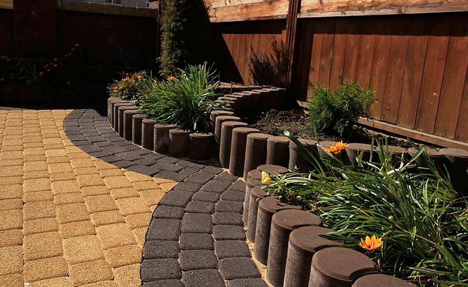 Ogród z dwukolorową ścieżką z kostki, obrzeża z betonowych palików, drewniane ogrodzenie w tle.