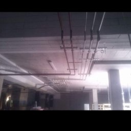 Mm Instal - Montaż instalacji rur pod sufitem w surowym wnętrzu, widoczne rury w kolorze miedzi i srebrnym, elementy mocujące, surowy betonowy sufit i słup konstrukcyjny.