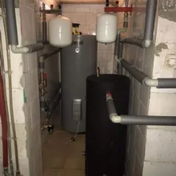 Kotłownia z dwoma zbiornikami akumulacyjnymi: szarym marki Ferroli i czarnym, oraz białymi naczyniami przeponowymi, instalacja rur w szarej izolacji, widok z wejścia do pomieszczenia.