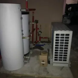 Zewnętrzna jednostka pompy ciepła powietrze-woda z podwójnym zbiornikiem buforowym, zainstalowana na betonowej posadzce, widoczne rury instalacyjne i karton obok urządzenia.