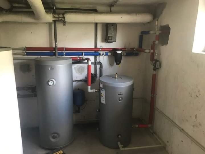 Dwa srebrne zbiorniki akumulacyjne w piwnicy, połączone rurami w kolorze czerwonym i niebieskim, z widocznymi elementami instalacji hydraulicznej na ścianie.