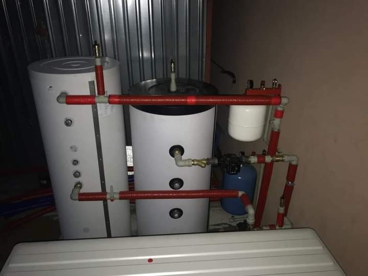 Dwa białe zbiorniki, połączone rurami w izolacji termicznej, z widocznymi elementami instalacji hydraulicznej i osprzętem, w pomieszczeniu technicznym.