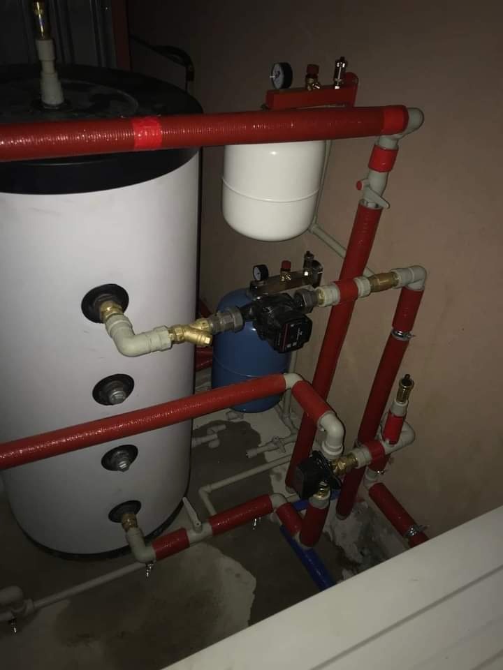 Biały zbiornik akumulacyjny z czerwonymi rurami izolowanymi termicznie, niebieski zbiornik przeponowy i liczne zawory w kotłowni.