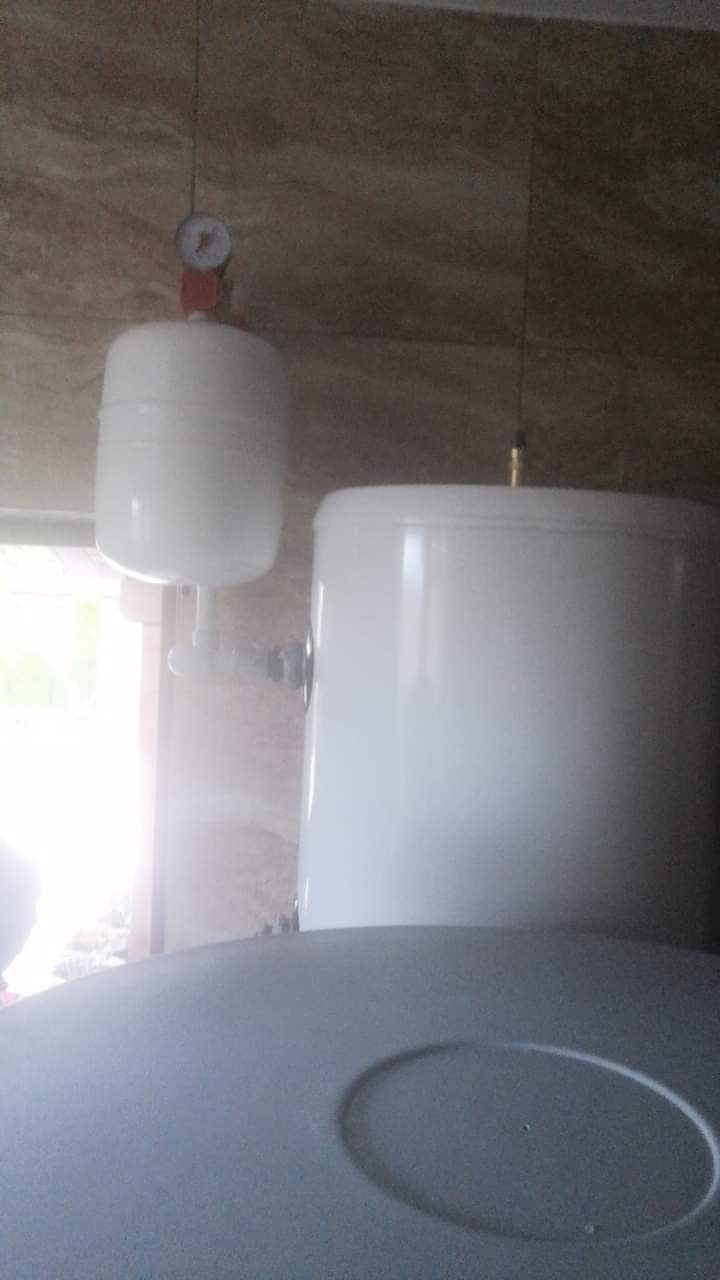 Dwa białe zbiorniki wyrównawcze w kotłowni, jeden z manometrem, zamontowane na tle płytek imitujących kamień.