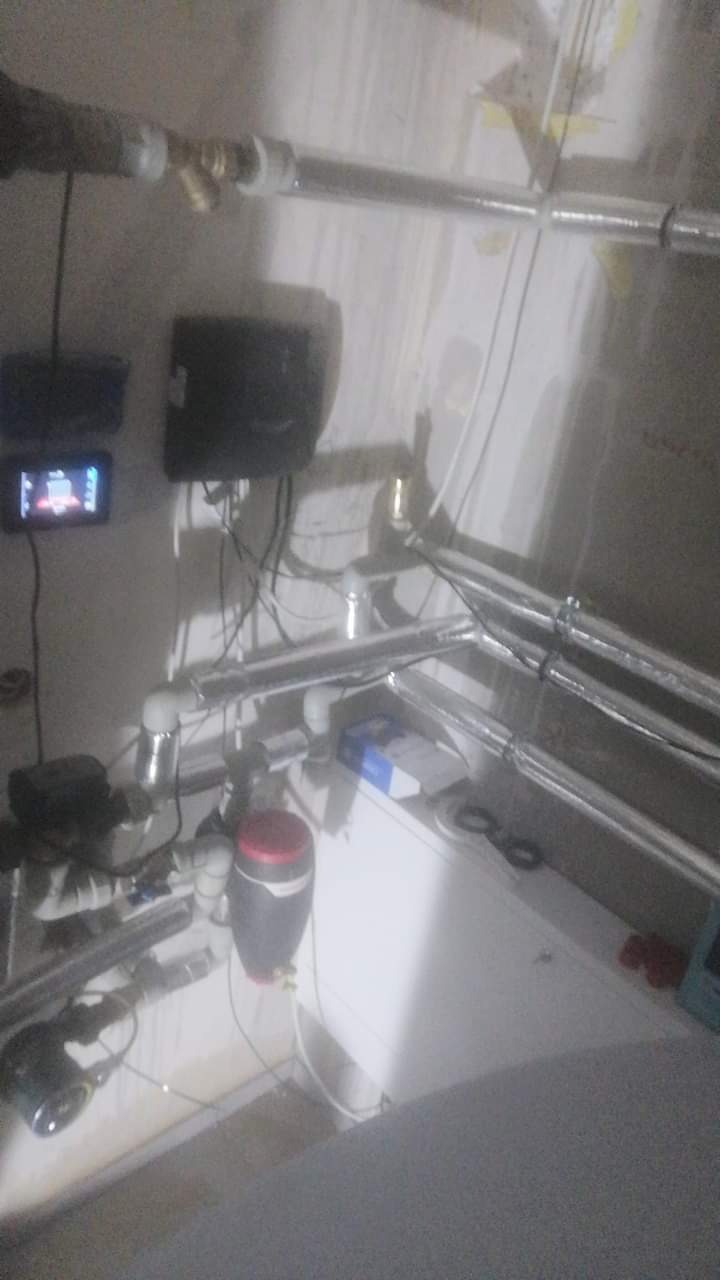 Fragment instalacji hydraulicznej z rurami w izolacji, zaworami, pompą obiegową z czerwonym elementem i sterownikiem na ścianie w surowym pomieszczeniu.