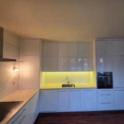 Paweł Goliński MNW - Nowoczesna, biała zabudowa kuchenna z błyszczącymi frontami, oświetleniem podszafkowym i drewnianą podłogą w mieszkaniu.