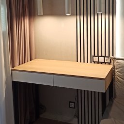 Paweł Goliński MNW - Biurko z jasnym drewnianym blatem i białymi szufladami, wkomponowane w aranżację sypialni z pionowymi panelami ściennymi i wiszącymi lampami w stylu industrialnym.