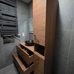 Paweł Goliński MNW - Drewniana szafka łazienkowa z otwartymi szufladami i czarną umywalką na blacie, na tle szarych płytek i grzejnika drabinkowego