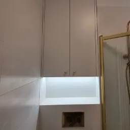 Biała szafka łazienkowa pod sufitem z oświetleniem LED nad złotym przyciskiem spłuczki, obok kabiny prysznicowej ze złotymi ramami.