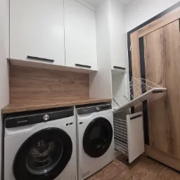 Funkcjonalna zabudowa pralni: białe szafki z czarnymi uchwytami, drewniany blat, dwie pralki Samsung i wysuwana suszarka w szafce. Drewniana podłoga.