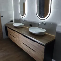 Nowoczesna łazienka z drewnianą szafką pod umywalki, owalnymi lustrami z podświetleniem LED i szarą płytką na ścianach.