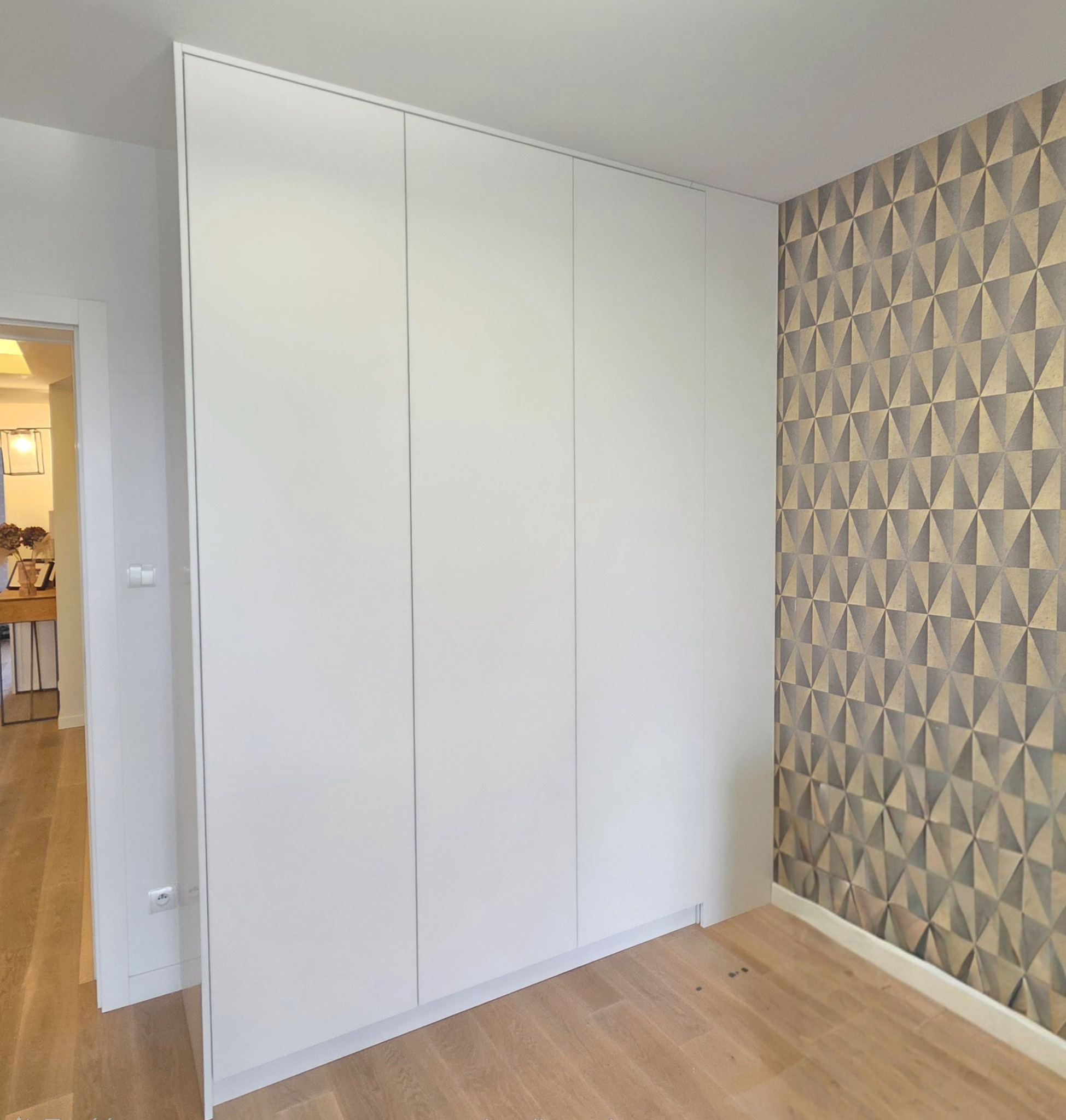 Biała, minimalistyczna szafa wnękowa z trzema drzwiami, przylegająca do ściany z geometryczną tapetą w odcieniach szarości i złota. Widoczna drewniana podłoga i fragment korytarza z oświetleniem.
