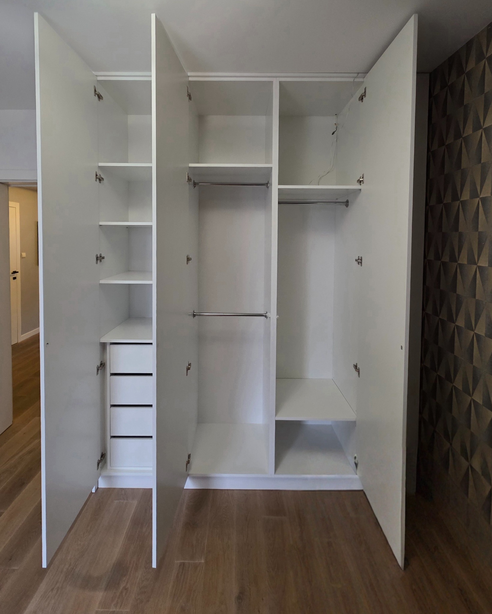 Biała szafa wnękowa z otwartymi drzwiami, widoczne półki, szuflady i drążki na ubrania, drewniana podłoga i wzorzysta tapeta w tle.