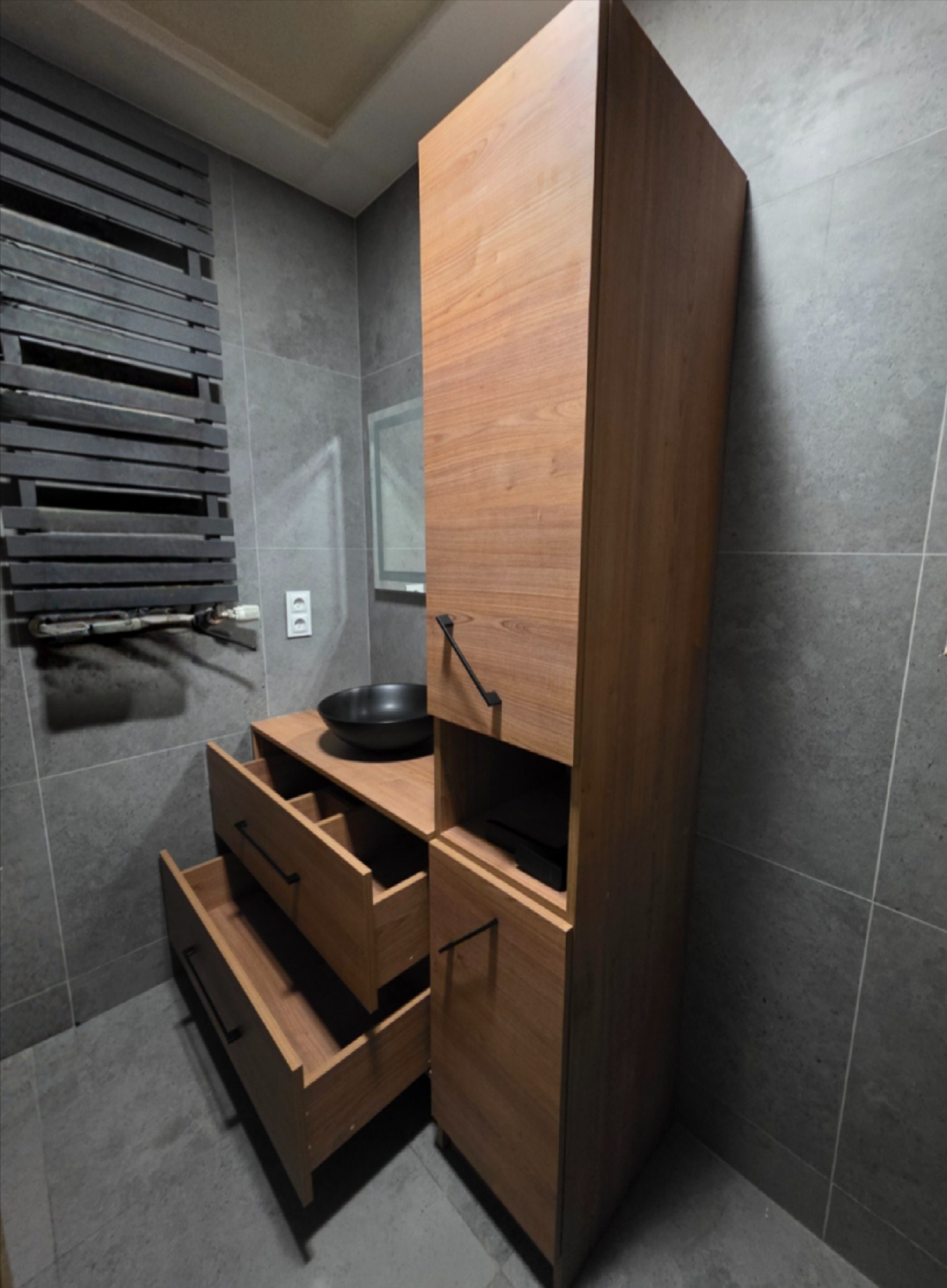 Drewniana szafka łazienkowa z otwartymi szufladami i czarną umywalką na blacie, na tle szarych płytek i grzejnika drabinkowego
