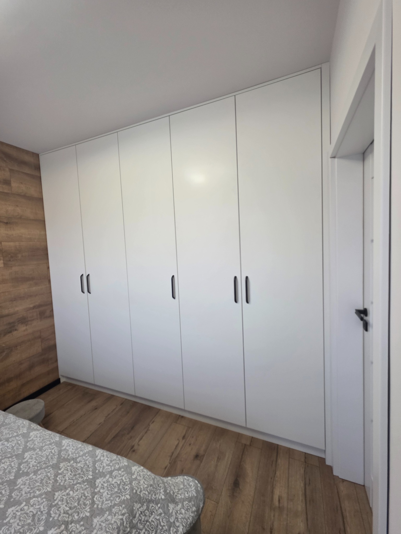 Biała szafa wnękowa z czarnymi uchwytami na tle drewnianej podłogi i ściany imitującej drewno, widok z boku łóżka z szarą pościelą w kwiaty.